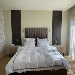 Nowoczesna sypialnia z łóżkiem małżeńskim w białej pościeli z ciemnymi paskami, wezgłowie z ciemnego weluru, symetryczne panele dekoracyjne na ścianie, lampki nocne oraz szafki w jasnym drewnie...