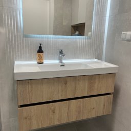 BDM Furniture - Nowoczesna łazienka z szafką pod umywalką w kolorze jasnego drewna, białą umywalką i podświetlanym lustrem; na blacie stoi butelka z dozownikiem.