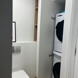 BDM Furniture - Pralka i suszarka w białej szafie na wymiar w łazience, widoczny fragment toalety i półki z jasnego drewna.