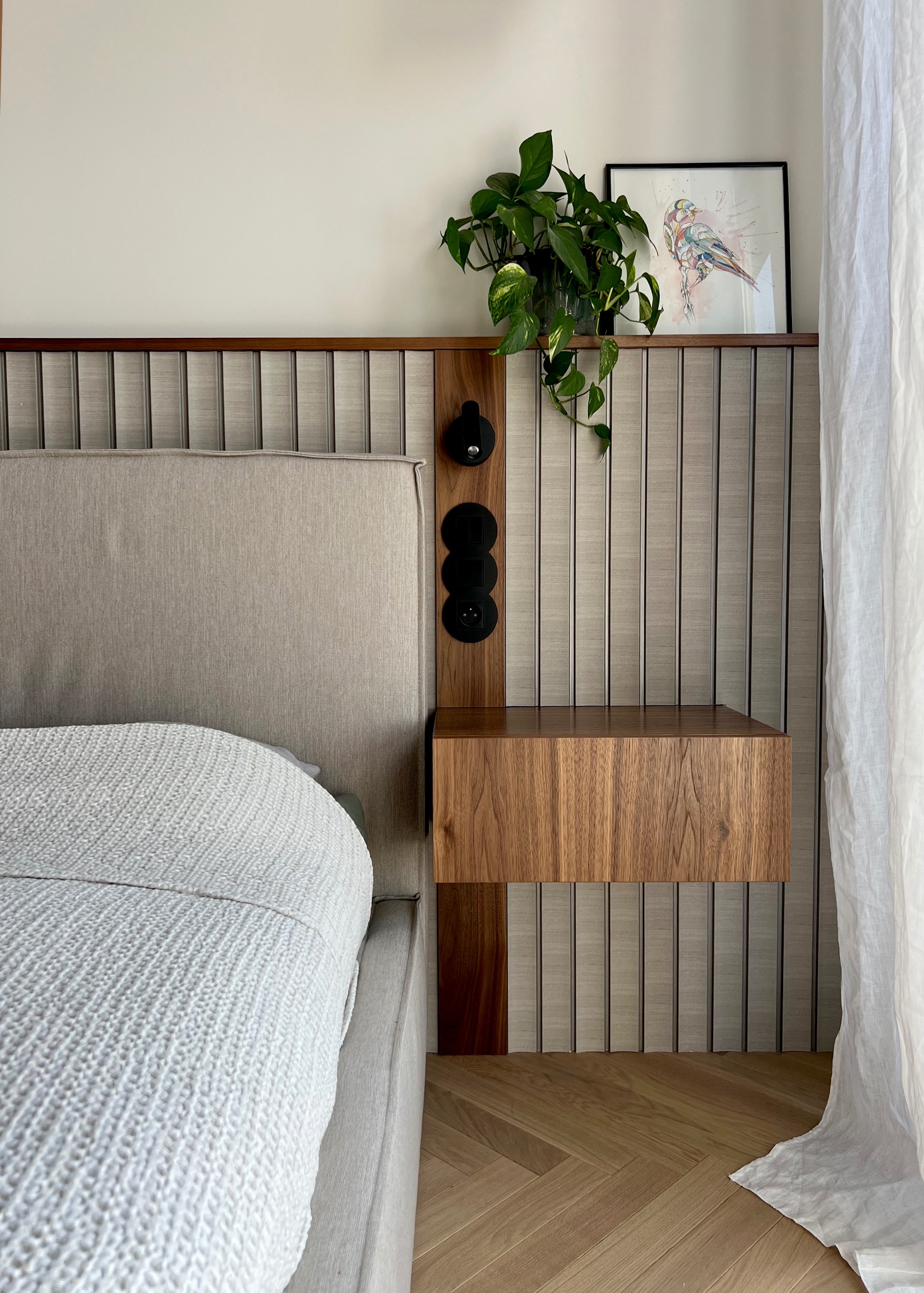 Nowoczesna sypialnia z łóżkiem z szarym zagłówkiem, drewnianą szafką nocną wiszącą z gniazdkami elektrycznymi, rośliną doniczkową i obrazkiem ptaka na ścianie z panelami dekoracyjnymi.