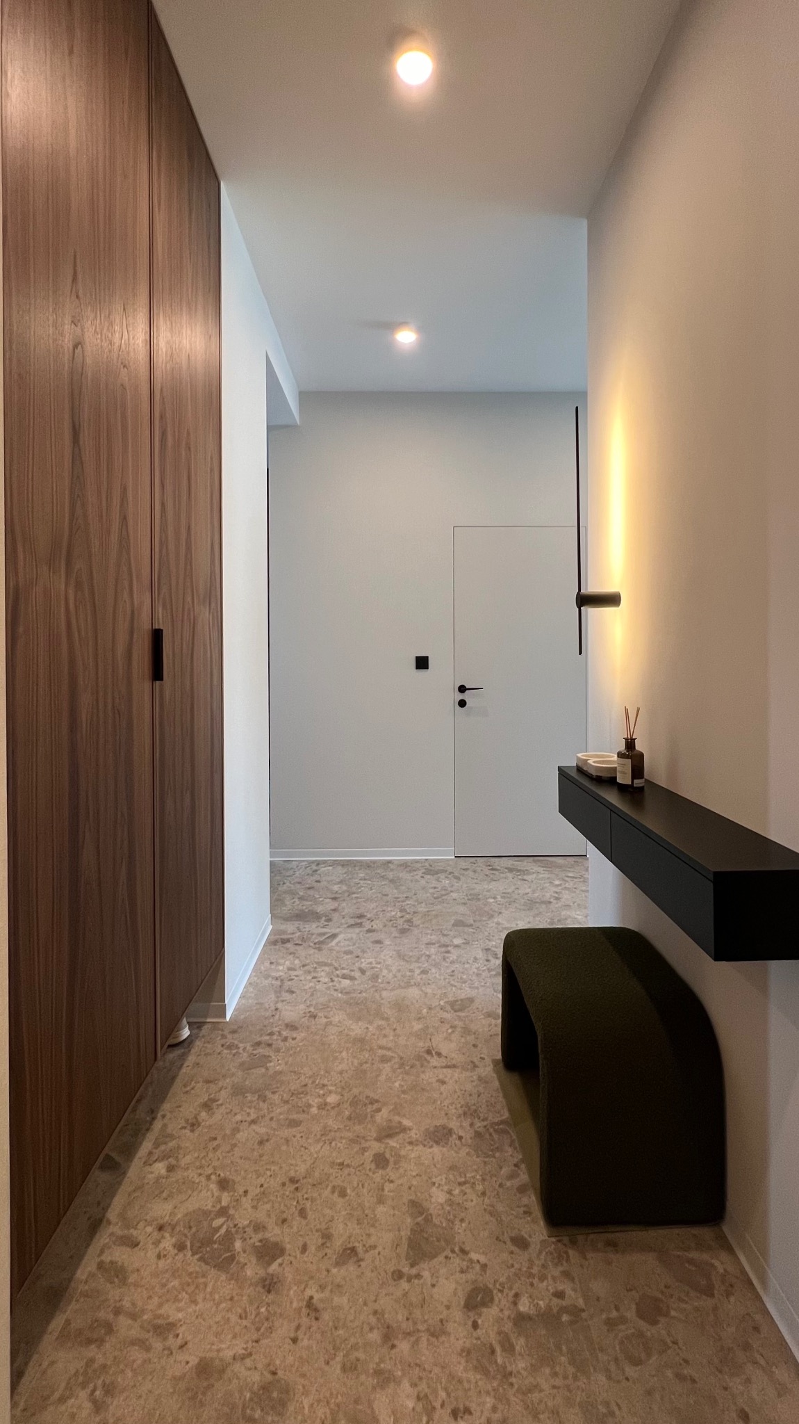 Wnętrze przedpokoju z dużą, drewnianą szafą, konsolą ścienną z dekoracjami, pufą obitą zielonym materiałem i podłogą z płytek imitujących kamień.