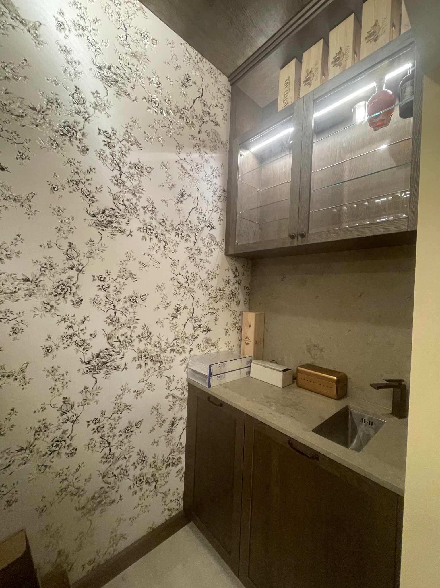 Niewielka kuchnia z ciemnymi szafkami, blatem z szarego kamienia i wzorzystą tapetą w kwiaty. Górne szafki z przeszklonymi drzwiczkami podświetlone LED, na górze drewniane skrzynki.