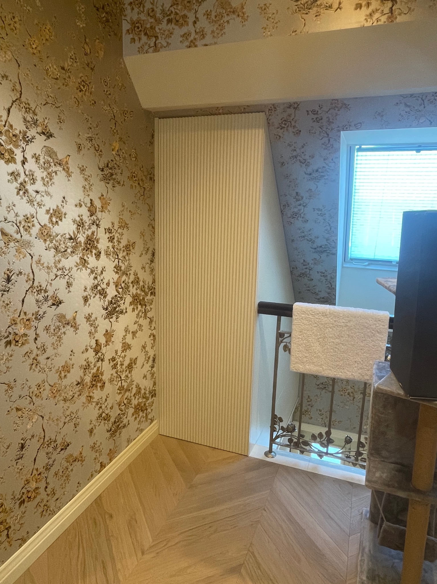 Wysoka, wąska szafa wnękowa z ryflowanymi drzwiami, wkomponowana w przestrzeń pod skosem, obok ściany pokrytej tapetą w kwiaty, podłoga w jasną jodełkę.