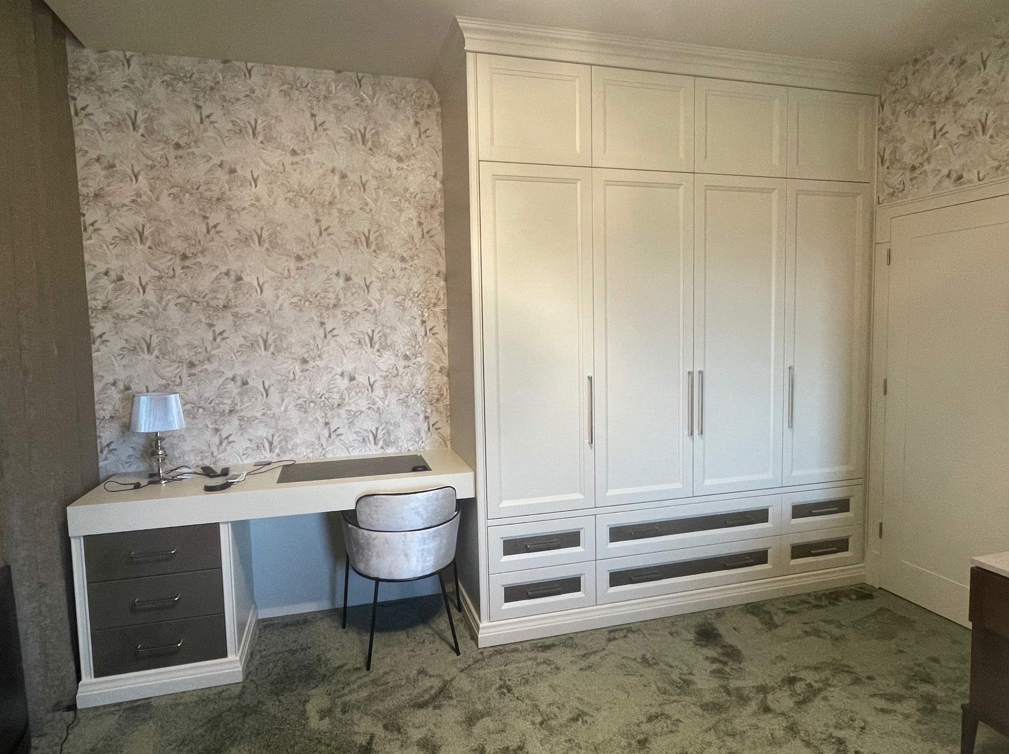 Biała szafa wnękowa z szufladami i biurkiem wbudowanym po lewej stronie, tapeta w tle z motywem roślinnym, srebrna lampka na biurku.