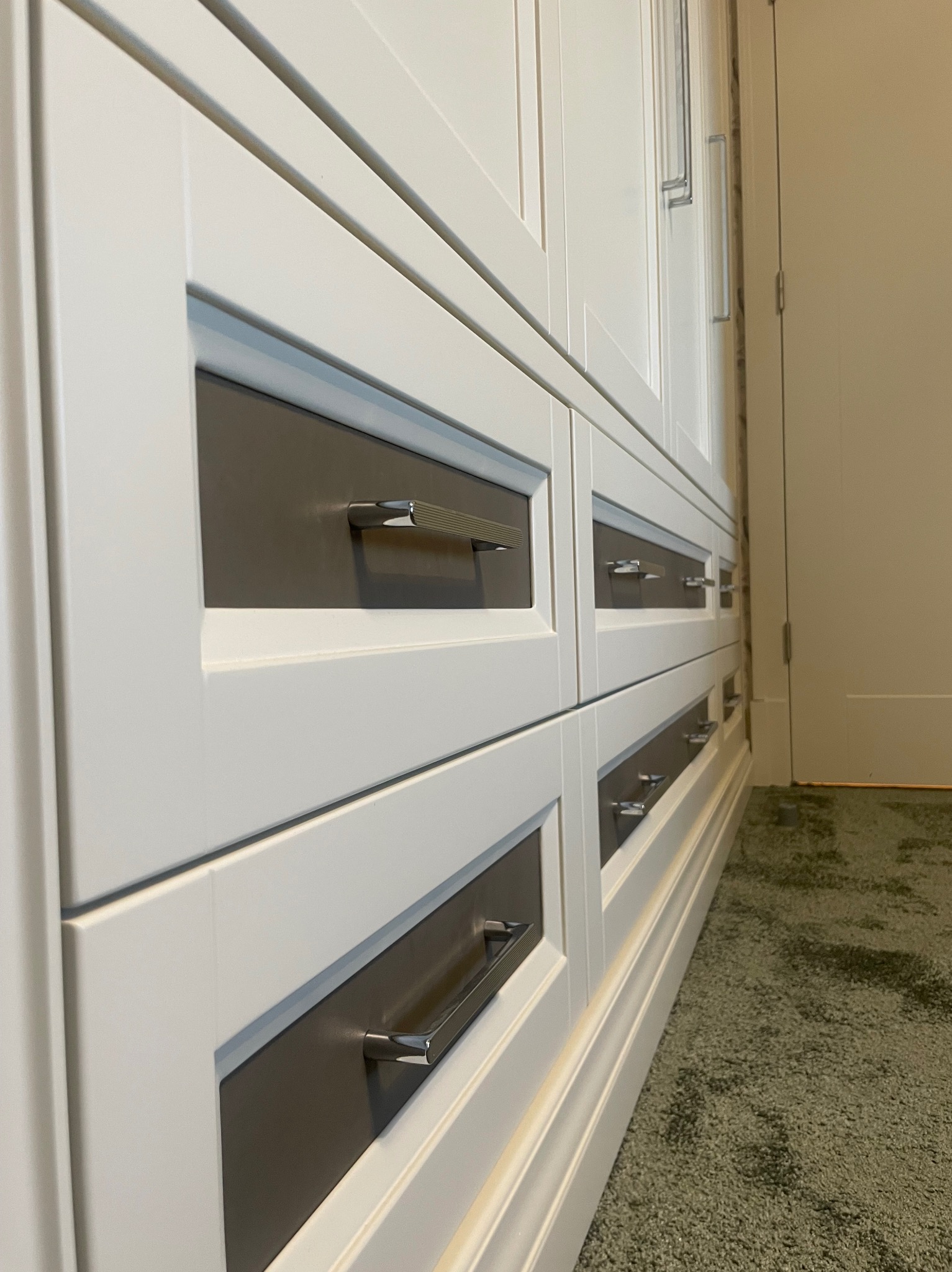 Biała szafa z szufladami i metalowymi uchwytami, widok z perspektywy podłogi na tle zielonego dywanu i białych drzwi.