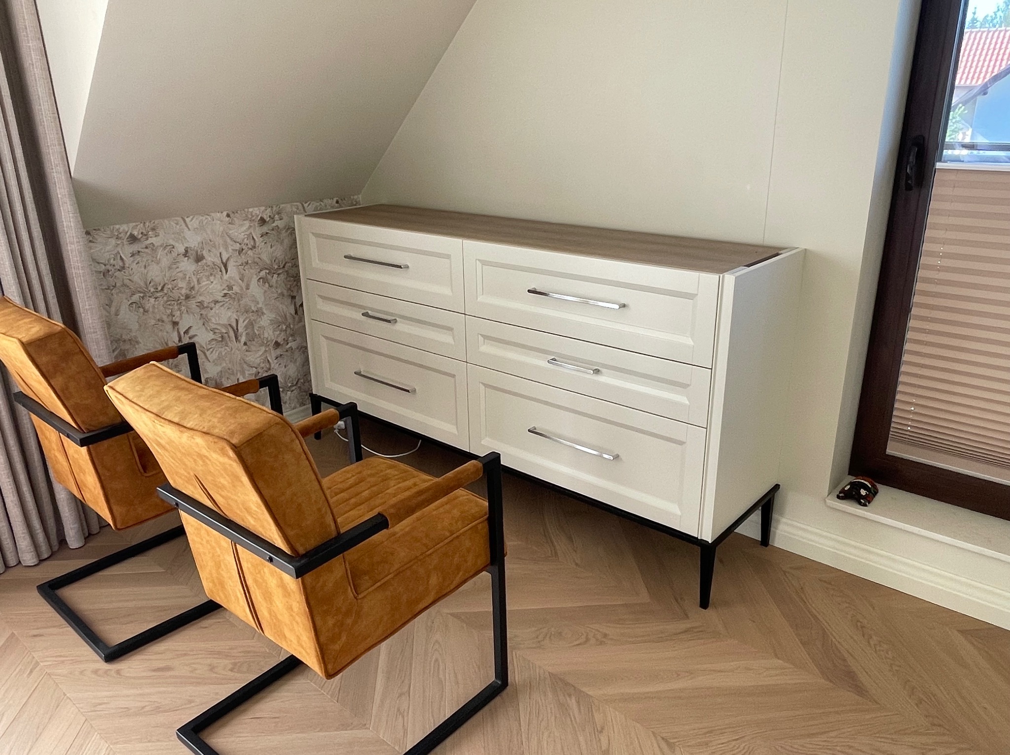 Biała komoda z sześcioma szufladami na czarnych nóżkach, z drewnianym blatem, stojąca w pokoju na poddaszu, obok dwa pomarańczowe fotele z czarną ramą, podłoga ułożona w jodełkę.