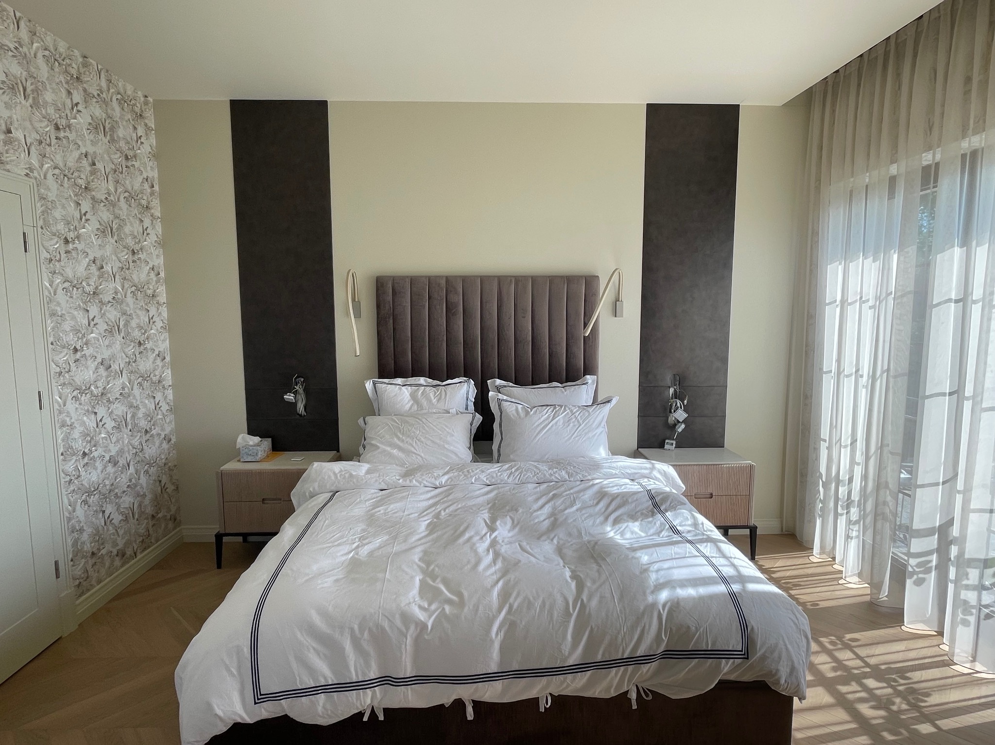 Nowoczesna sypialnia z łóżkiem małżeńskim w białej pościeli z ciemnymi paskami, wezgłowie z ciemnego weluru, symetryczne panele dekoracyjne na ścianie, lampki nocne oraz szafki w jasnym drewnie...