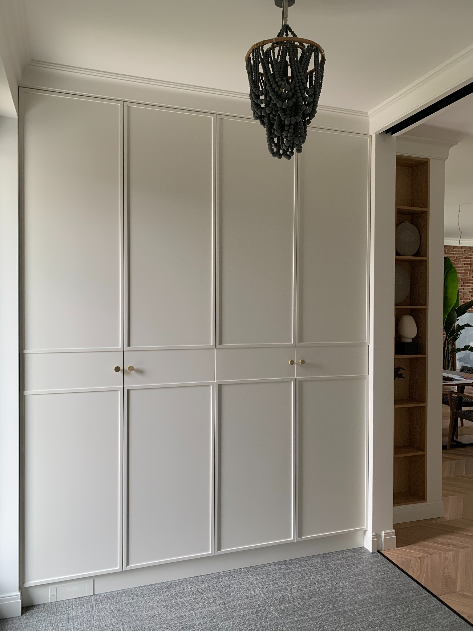 Elegancka, biała szafa wnękowa z dekoracyjnymi listwami i złotymi gałkami w Mszczonowie, uzupełniona oryginalnym, ciemnym żyrandolem z drewnianych koralików.