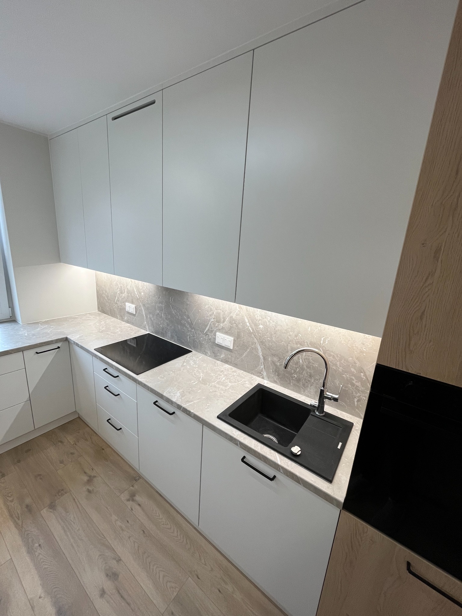 Nowoczesna kuchnia z białymi szafkami, blatem imitującym marmur, czarnym zlewem i płytą indukcyjną, oświetlona ledami pod szafkami wiszącymi.