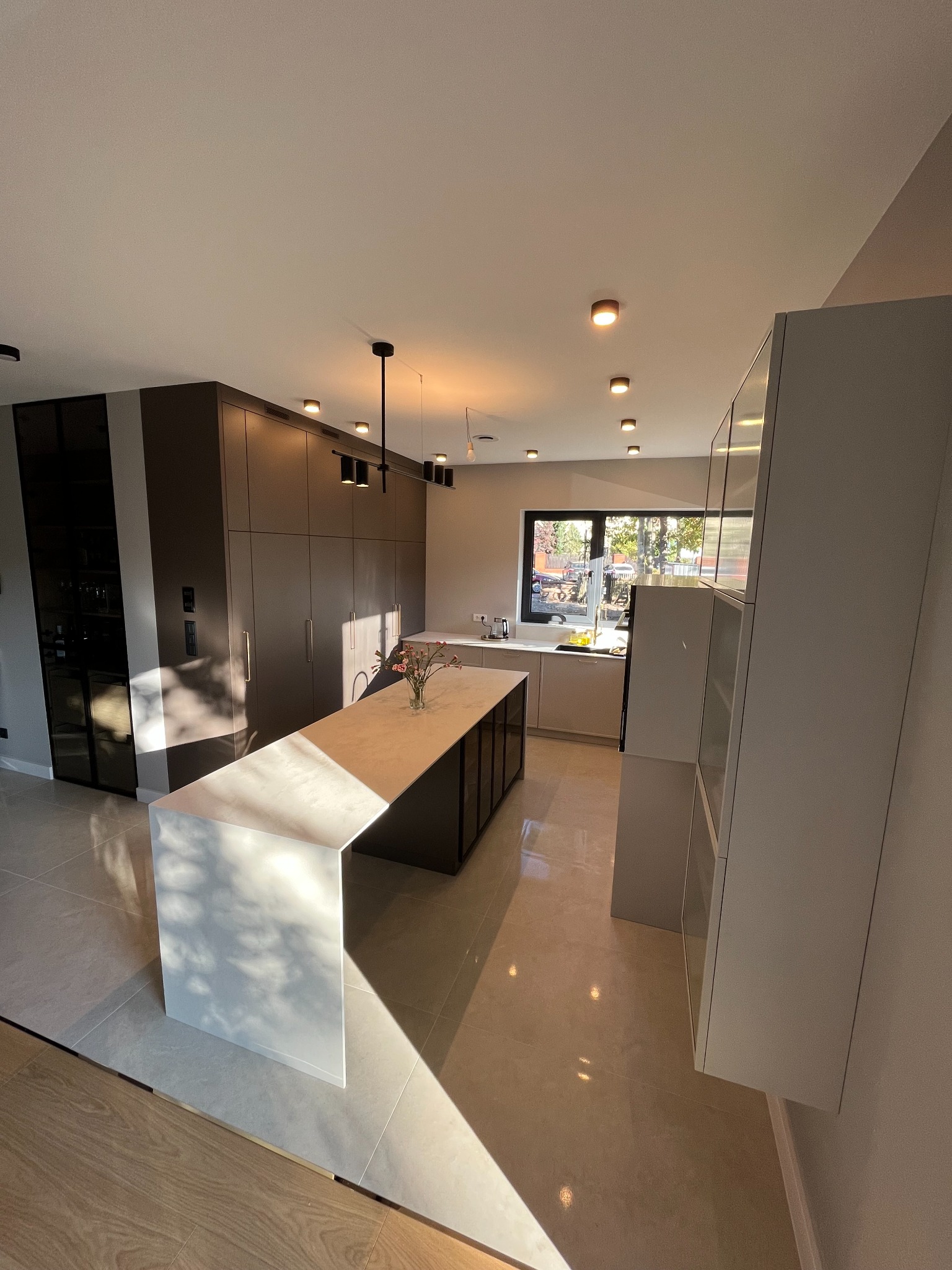 Nowoczesna kuchnia z wyspą i dużymi szafami w zabudowie, oświetlona ciepłym światłem słonecznym padającym przez okno.