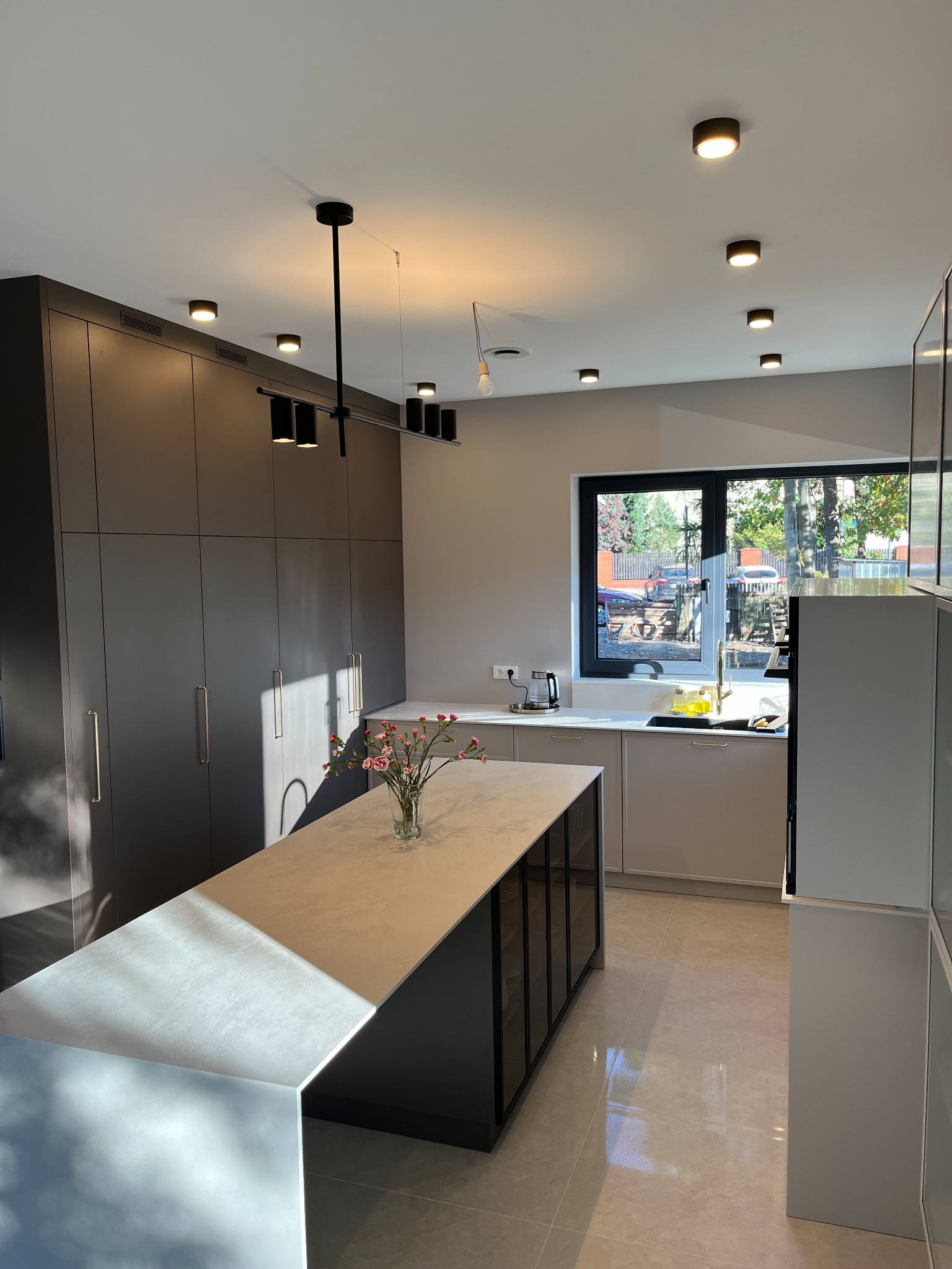 Nowoczesna kuchnia w odcieniach szarości z wyspą i zabudową po sufit, oświetlona punktowymi lampami, z widokiem na zieleń za oknem. Na wyspie stoi wazon z kwiatami.