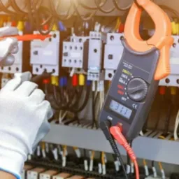 Elektryk w białych rękawicach ochronnych mierzy parametry elektryczne w rozdzielni za pomocą miernika cęgowego wskazującego 311, z widocznymi bezpiecznikami i przewodami.