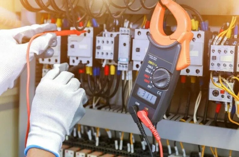 Elektryk w białych rękawicach ochronnych mierzy parametry elektryczne w rozdzielni za pomocą miernika cęgowego wskazującego 311, z widocznymi bezpiecznikami i przewodami.