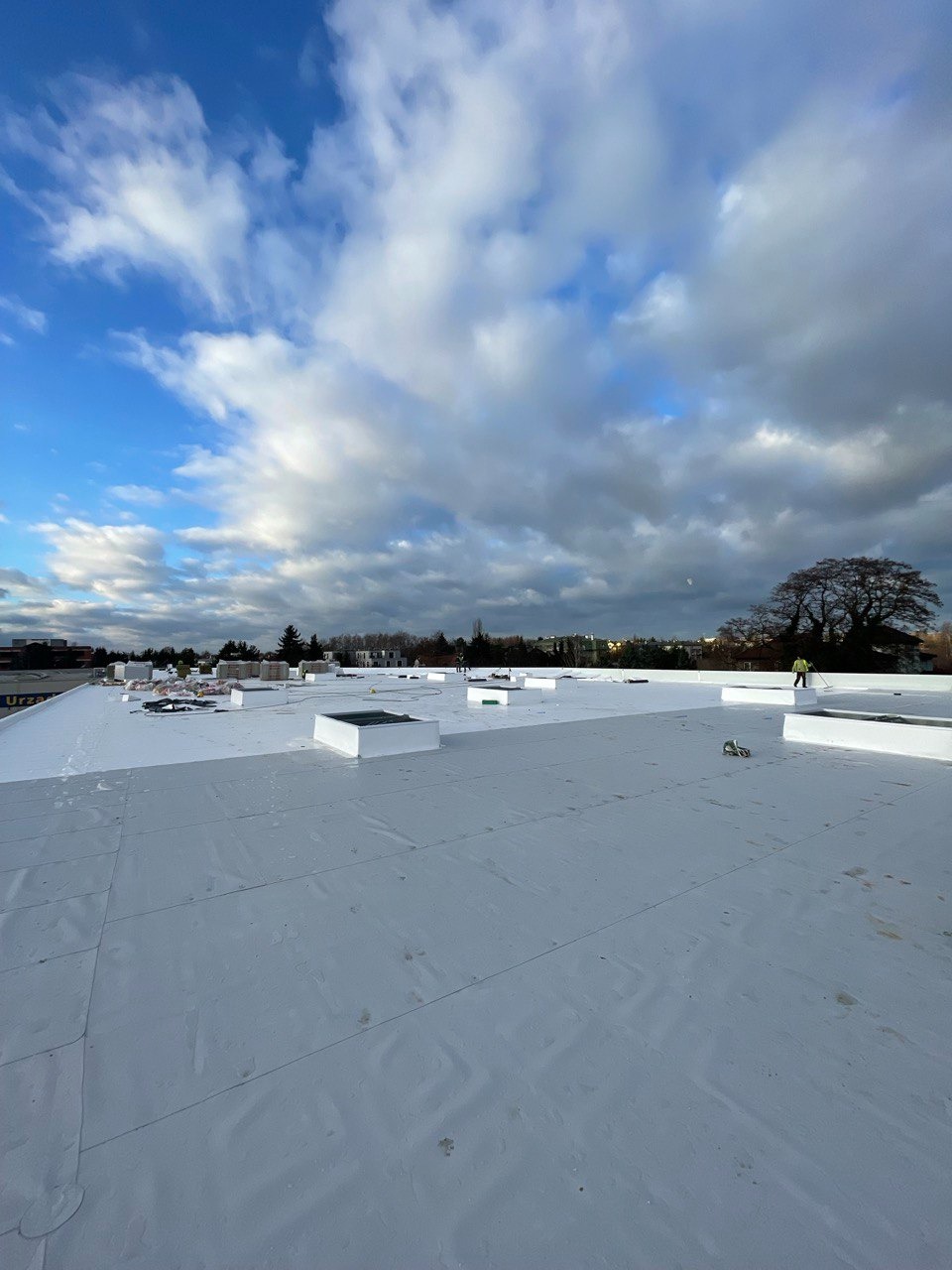 Biała, płaska powierzchnia dachu pokryta membraną, widoczne świetliki dachowe i pracownik w oddali, pochmurne niebo.