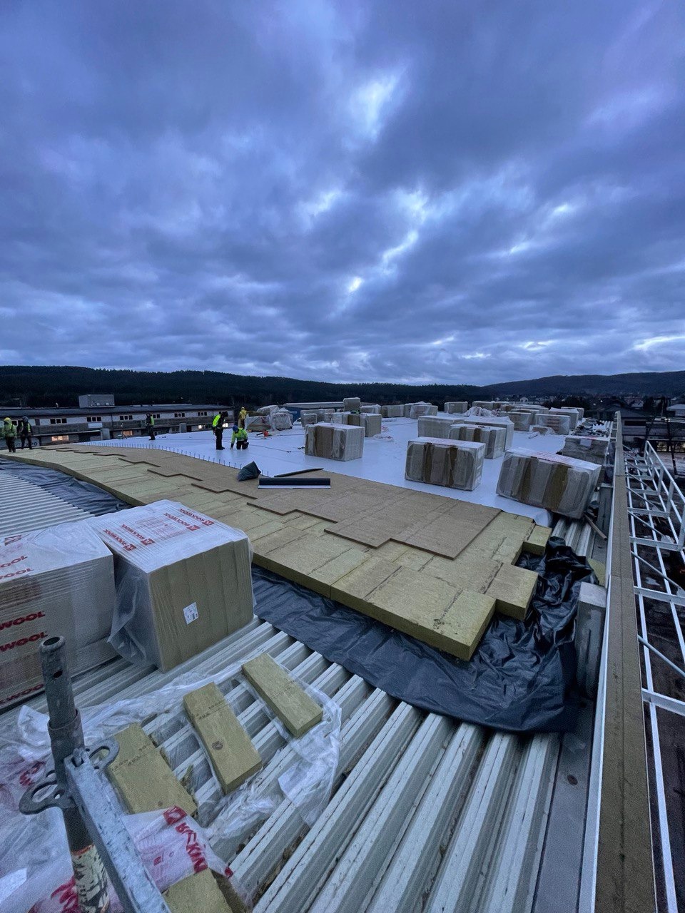 Montaż izolacji termicznej na dachu przemysłowym, widoczne płyty izolacyjne Rockwool, folia paroizolacyjna i ekipa pracowników na tle pochmurnego nieba.