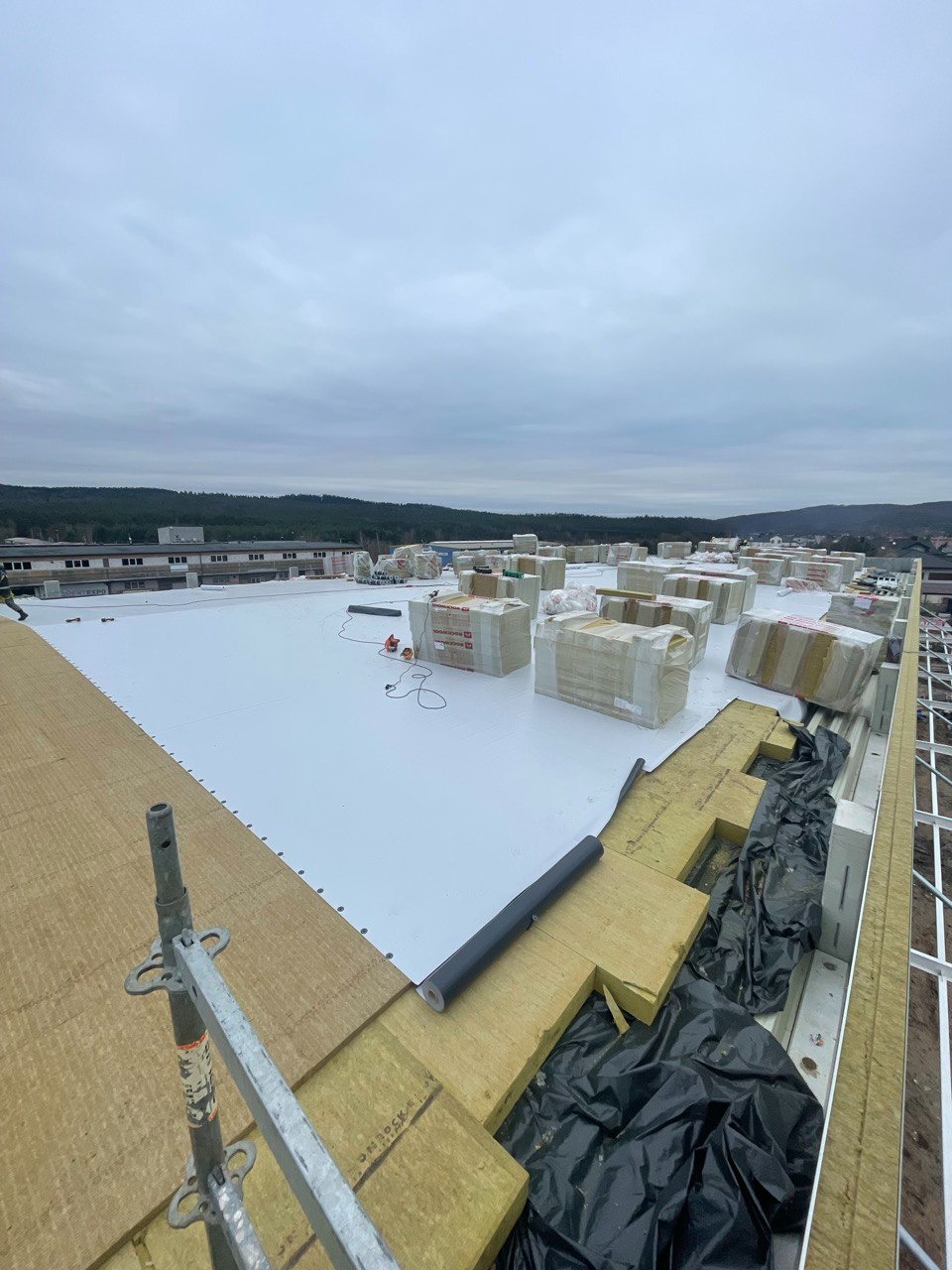 Montaż izolacji termicznej z wełny mineralnej na płaskim dachu przemysłowym, widoczne rolki papy i narzędzia, w tle zabudowania miejskie pod pochmurnym niebem.