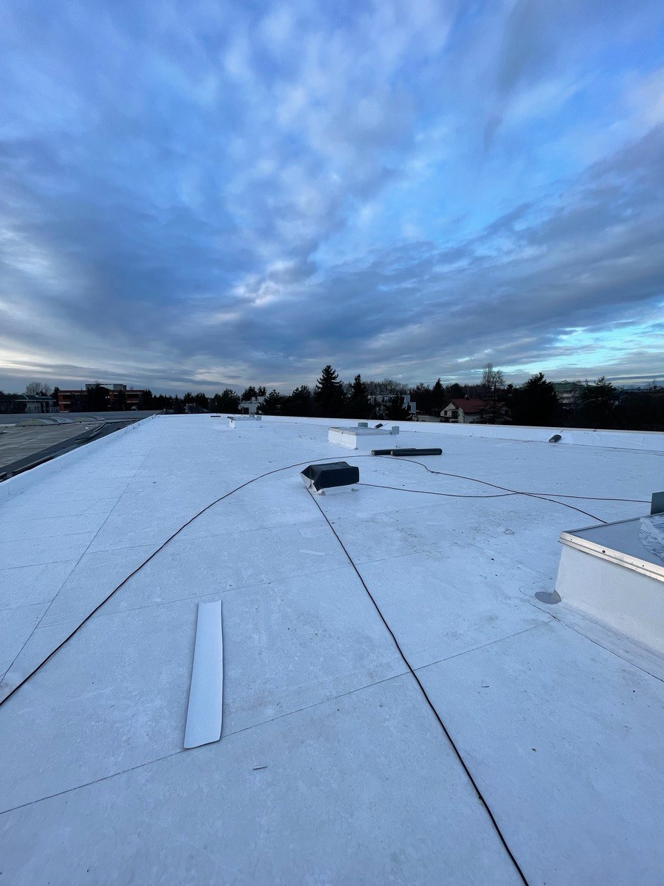 Widok z góry na płaski, biały dach komercyjny z kilkoma elementami wentylacyjnymi i przewodami, na tle pochmurnego nieba.