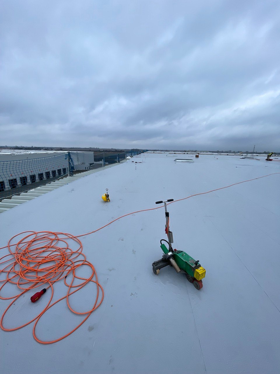 Zielony automat spawalniczy do membran dachowych na szarym, płaskim dachu przemysłowym, obok zwinięty pomarańczowy kabel i żółty kask, pochmurne niebo w tle.