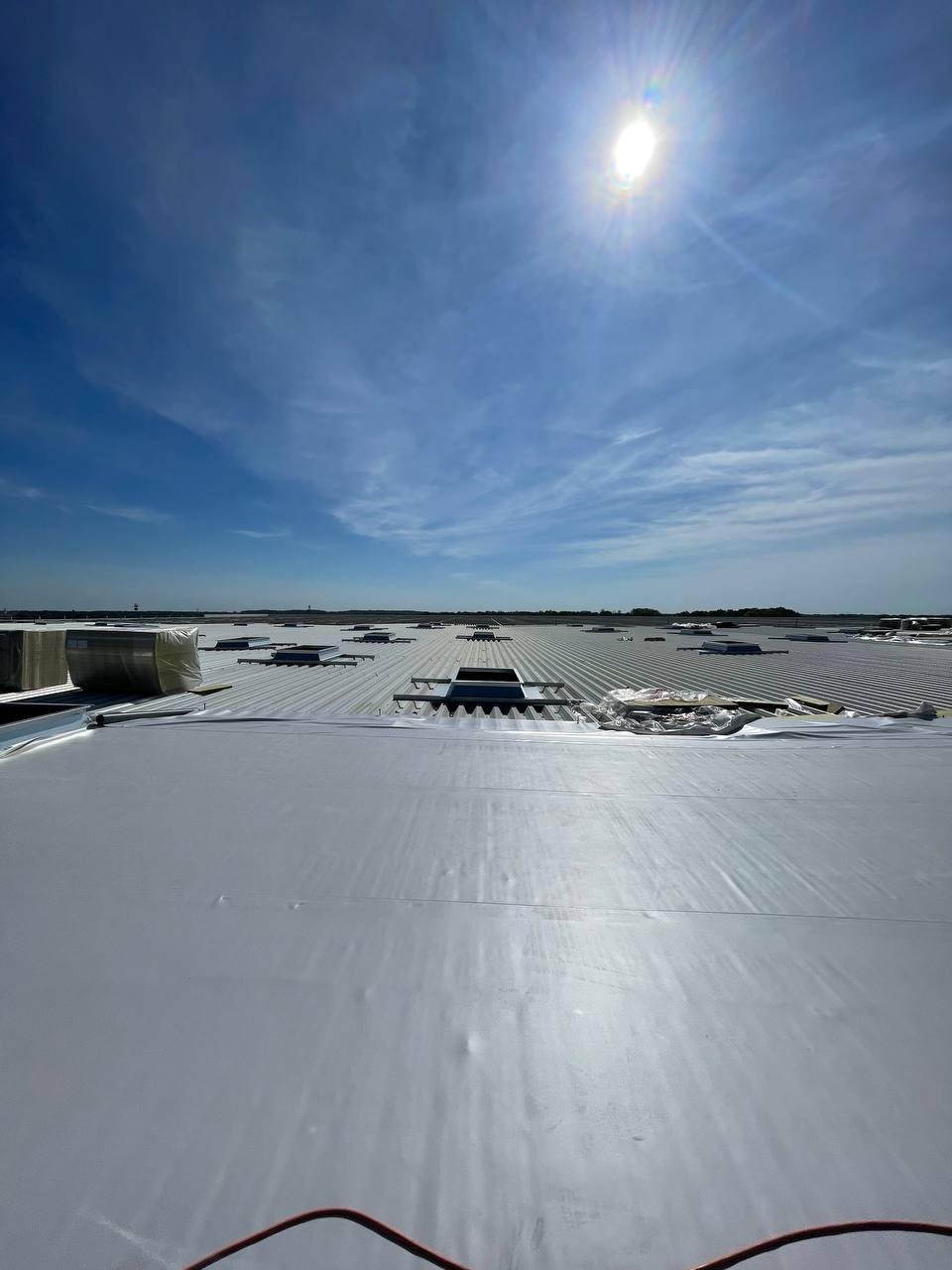 Szeroki kadr na membranę dachową z widocznymi świetlikami na dachu przemysłowym, jasne niebo z lekkimi chmurami i intensywnym słońcem w tle.