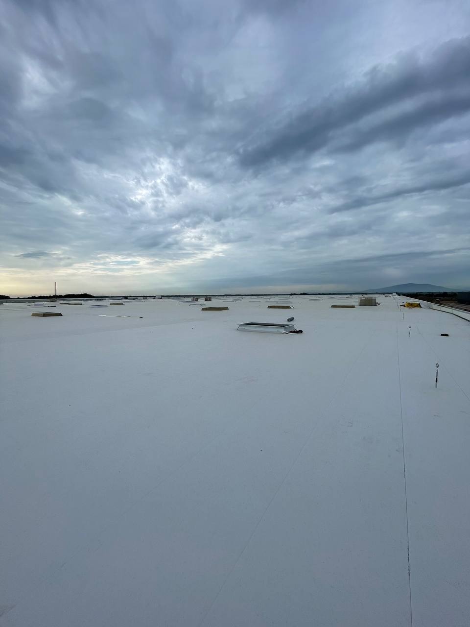 Widok z góry na płaski, biały dach przemysłowy z kilkoma świetlikami i antenami na tle pochmurnego nieba.