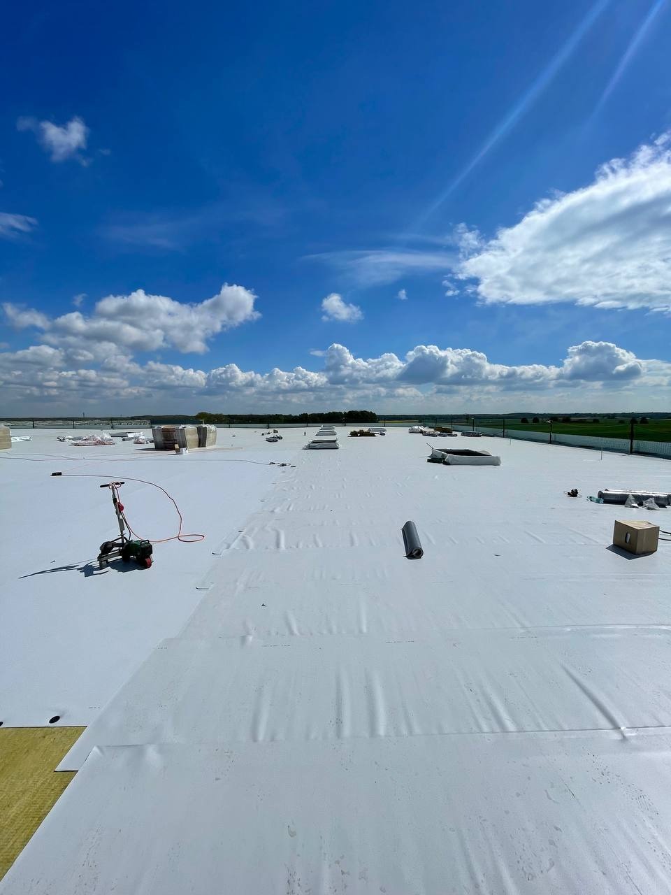 Biała membrana dachowa rozłożona na płaskim dachu, widoczna zgrzewarka do membran, błękitne niebo z chmurami w tle.