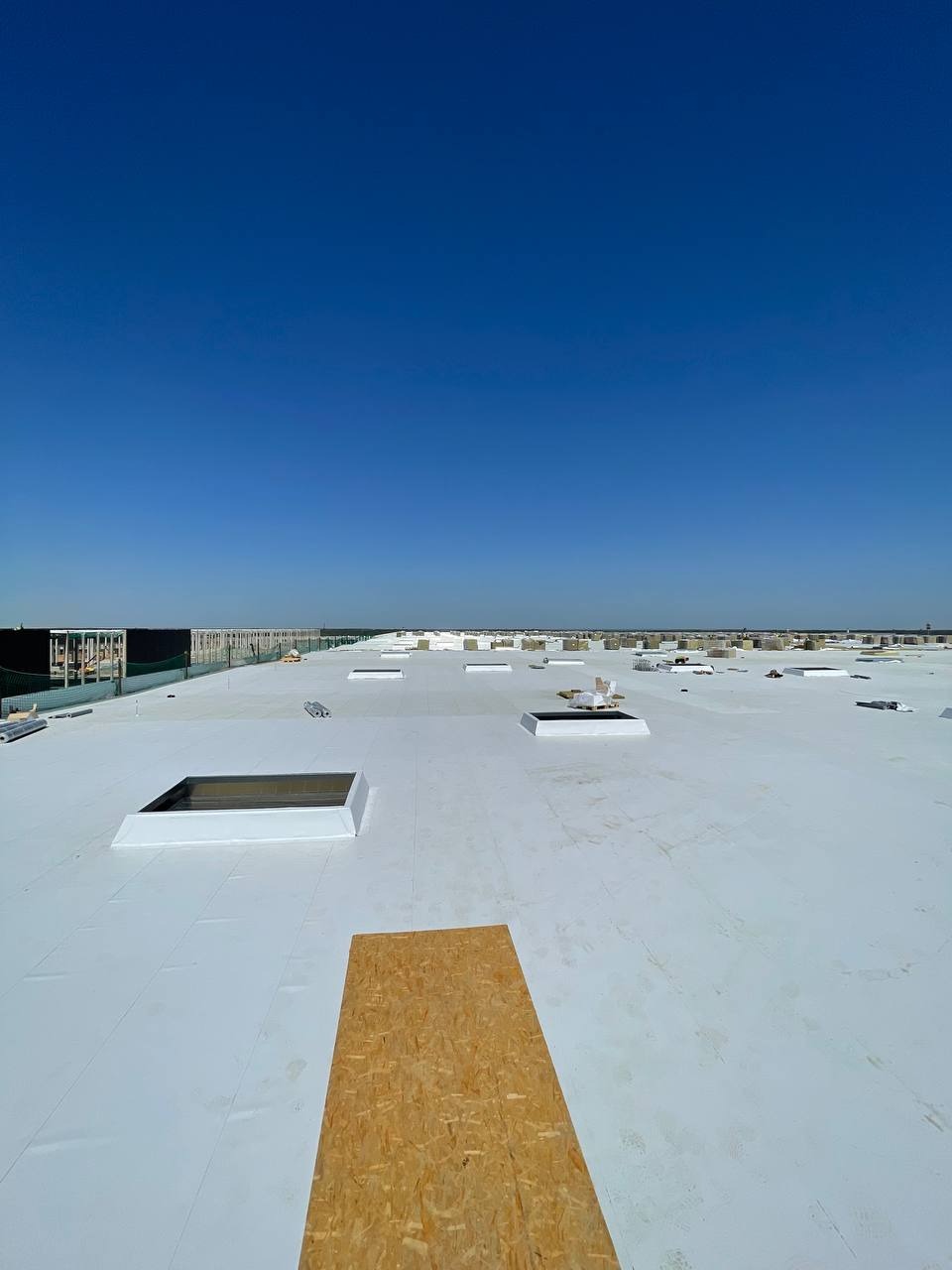 Biała membrana dachowa na płaskim dachu przemysłowym z świetlikami, deska OSB w pierwszym planie, błękitne niebo w tle.