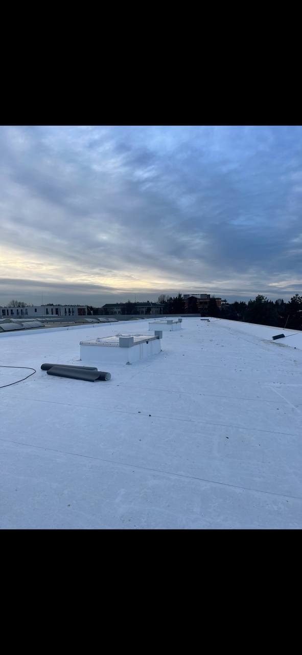 Biały, płaski dach budynku z widocznymi kominami wentylacyjnymi i rolkami papy, na tle pochmurnego nieba.