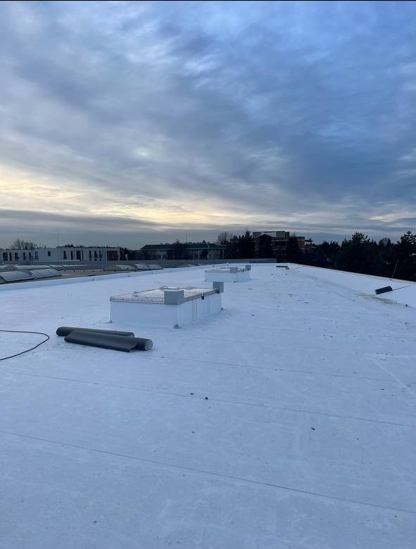 Płaski dach pokryty jasną membraną, widoczne kominki wentylacyjne i rolki materiału, budynki w tle pod pochmurnym niebem.
