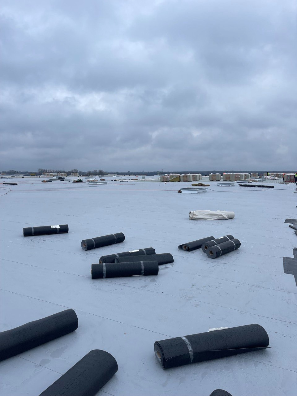 Rozwinięte rolki czarnej papy na płaskim dachu w trakcie prac, widoczne zapasy materiałów i robotnik w oddali, pochmurne niebo w tle.