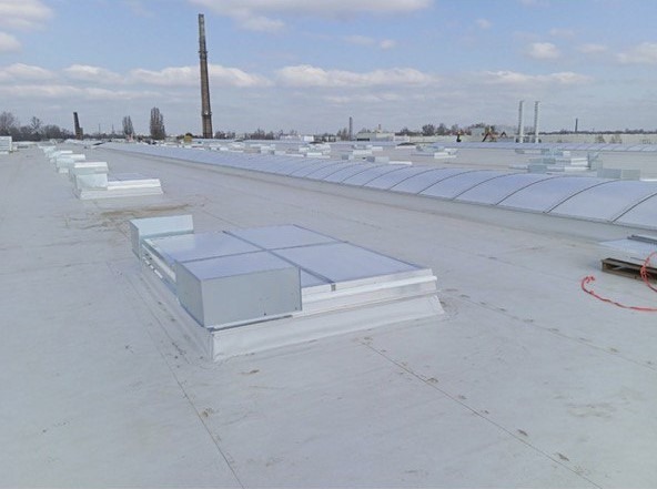 Biały dach przemysłowy z świetlikami dachowymi i komin fabryczny w tle, widok z góry.