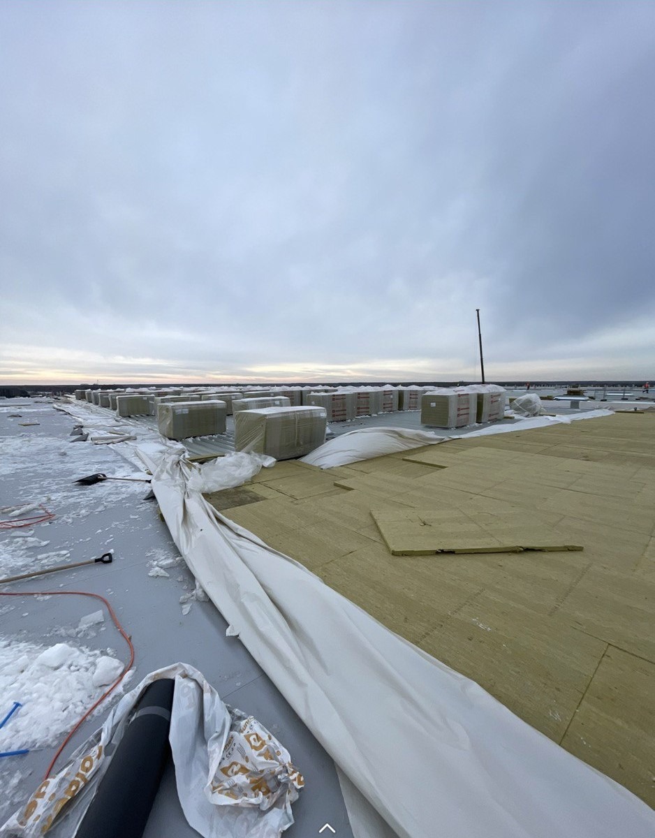 Płaski dach w trakcie prac: izolacja z wełny mineralnej, zabezpieczone palety z materiałem budowlanym, widoczny śnieg i pochmurne niebo.