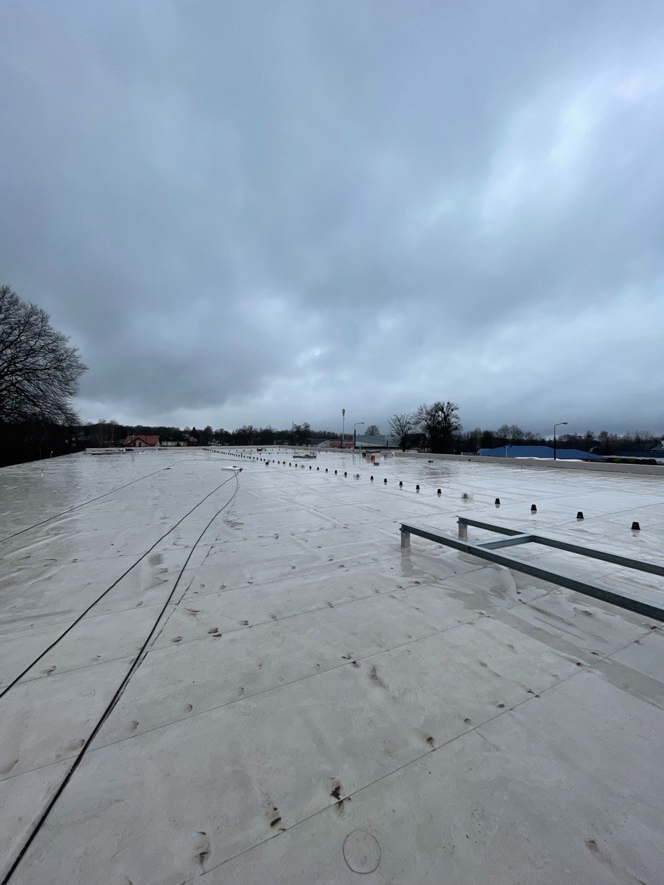 Biała, płaska powierzchnia dachu przemysłowego z widocznymi łączeniami paneli, elementami mocującymi i metalową konstrukcją pod instalacje, na tle pochmurnego nieba i odległych budynków.