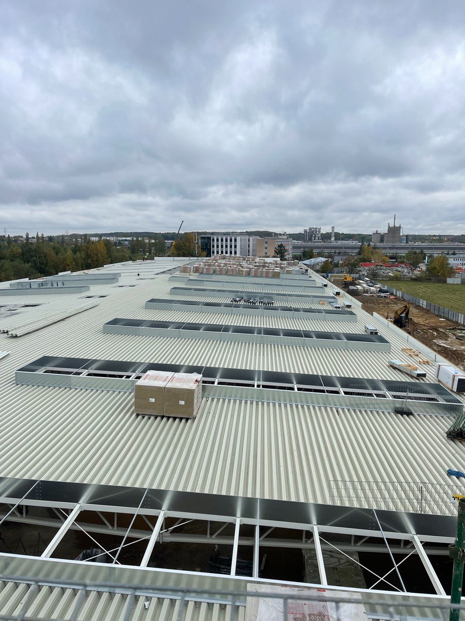Montaż blachy trapezowej na dachu hali przemysłowej, widoczne świetliki dachowe w metalowych obramowaniach, przygotowane materiały budowlane w kartonach. W tle zabudowa miejska i pochmurne niebo.