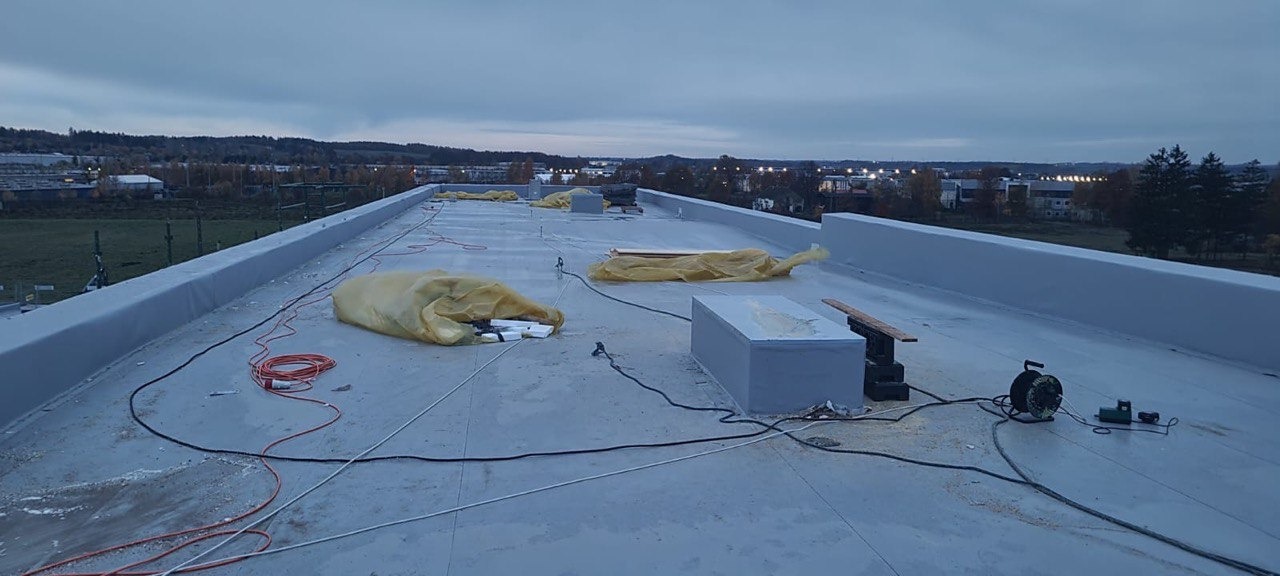Płaski dach budynku w trakcie prac, widoczne narzędzia, materiały zabezpieczone folią, oraz elementy instalacji na tle zabudowy miejskiej pod pochmurnym niebem.