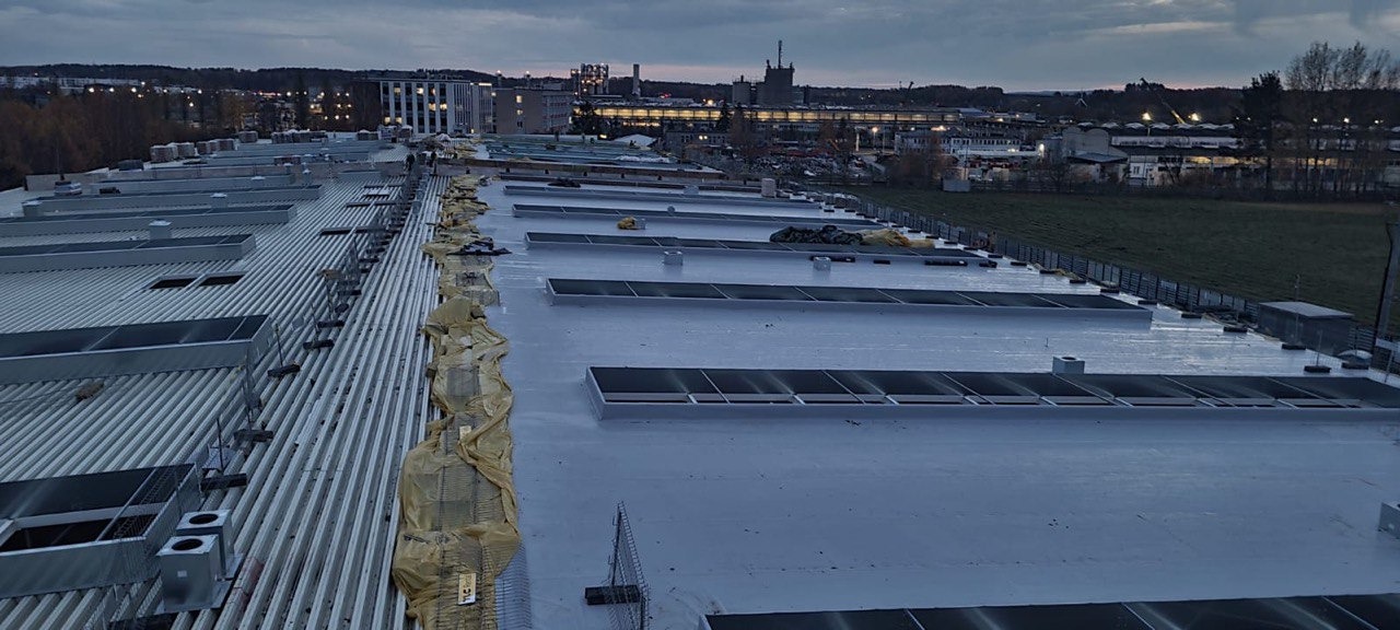 Szeroki widok na przemysłowy dach z blachy trapezowej, częściowo pokryty szarą powłoką, z widocznymi świetlikami i elementami instalacji wentylacyjnej, zabezpieczony żółtą folią wzdłuż krawędzi,...