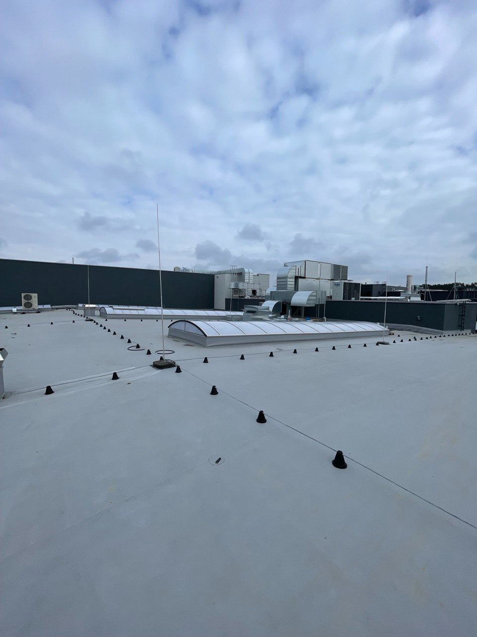 Jasnoszary dach przemysłowy z świetlikami kopułowymi i instalacją odgromową na tle pochmurnego nieba.