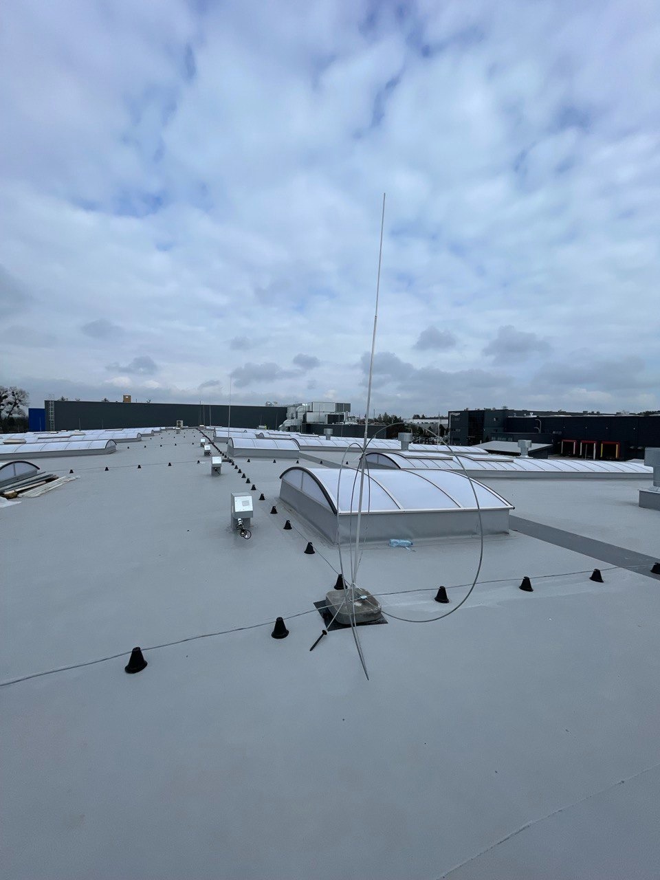 Płaski dach przemysłowy z szarym pokryciem, świetlikami kopułowymi, odgromnikami i elementami wentylacyjnymi, widok z góry przy pochmurnej pogodzie.