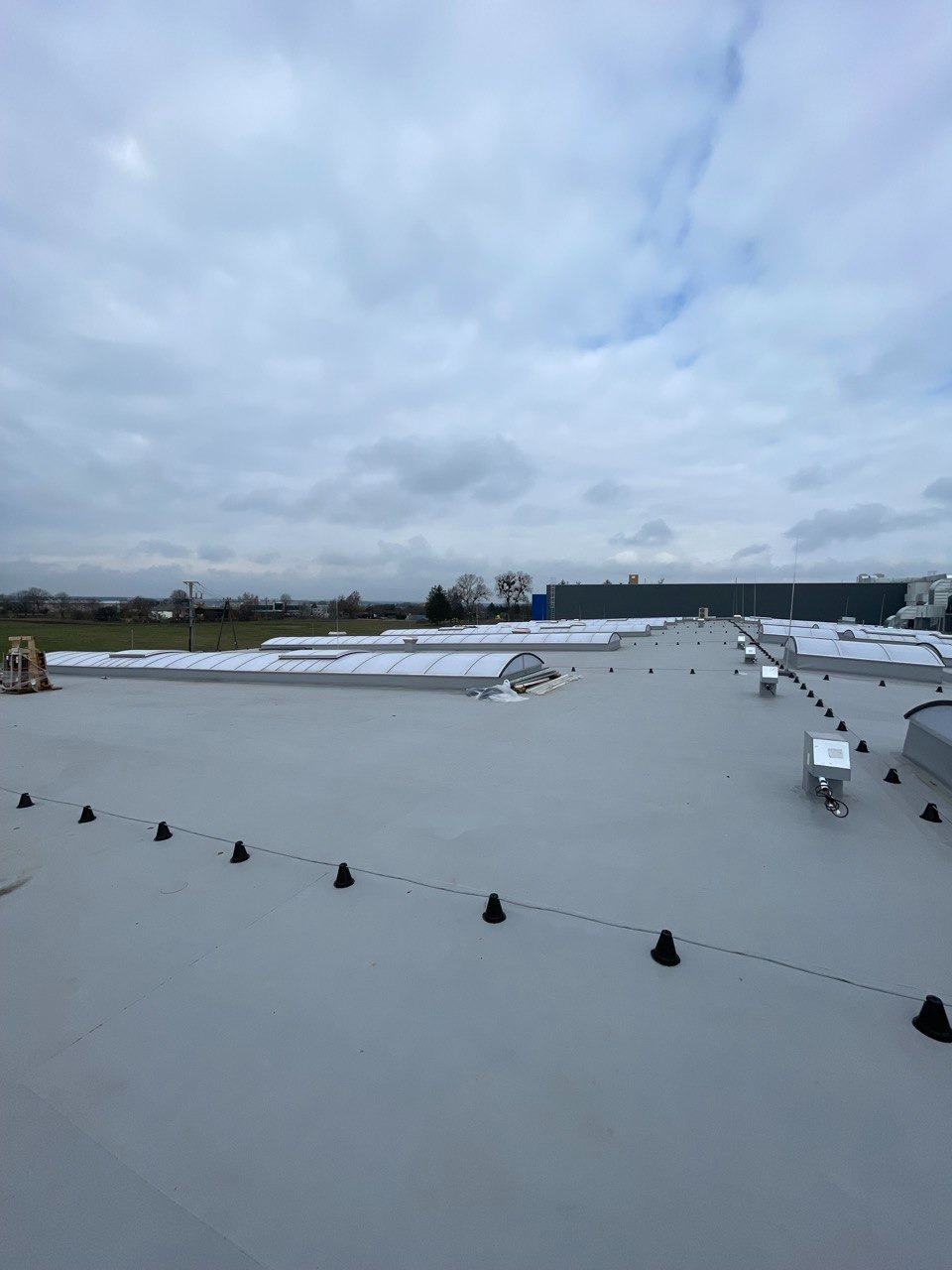 Widok z góry na płaski dach przemysłowy z świetlikami w Mińsku Mazowieckim, zabezpieczony systemem odgromowym z czarnymi uchwytami.