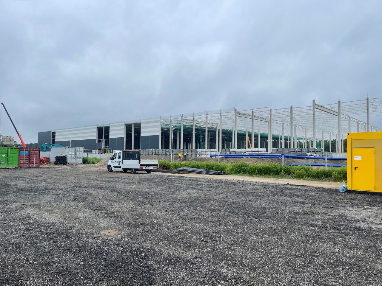 Budowa hali przemysłowej w trakcie realizacji, widoczna konstrukcja stalowa, częściowo obłożona płytami warstwowymi, na placu budowy kontenery i samochód dostawczy.