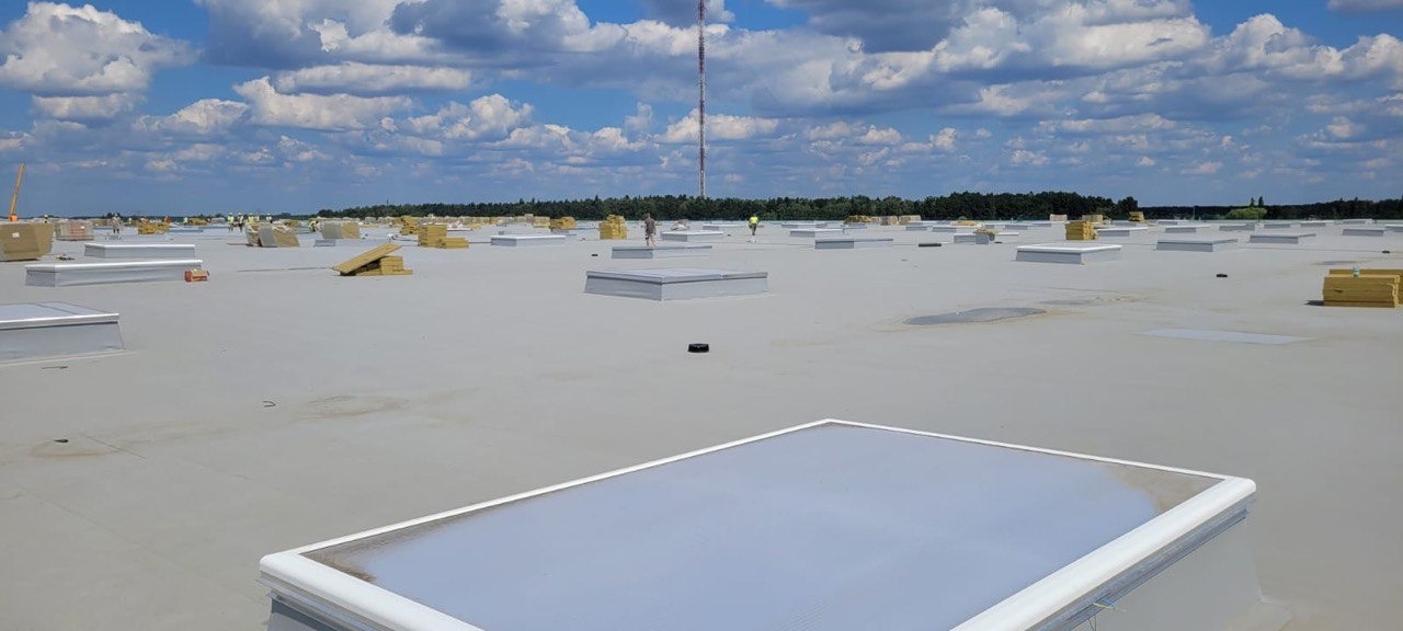 Rozległy, płaski dach przemysłowy z zamontowanymi świetlikami dachowymi, widoczne materiały budowlane i pracownicy w oddali, błękitne niebo z chmurami.