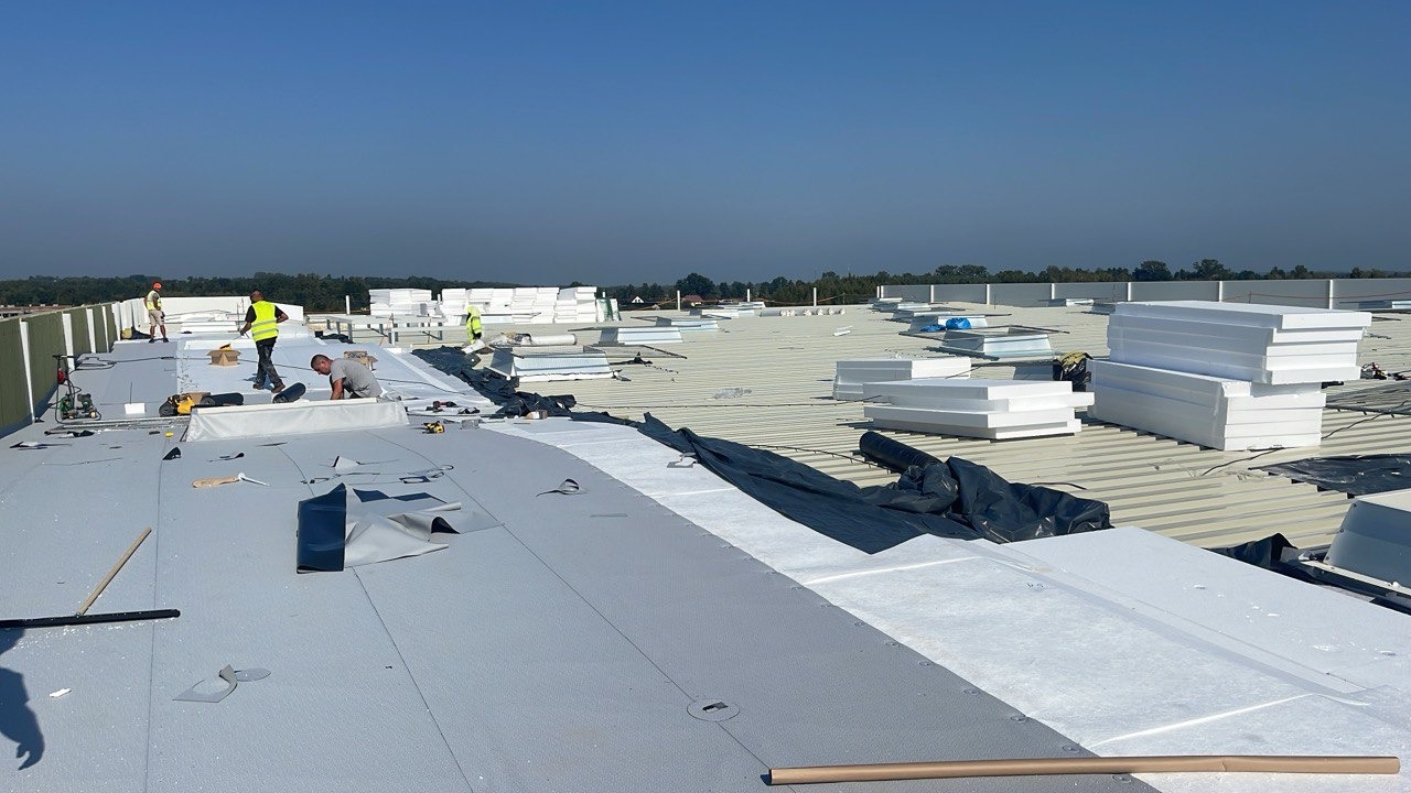 Prace na dachu przemysłowym: robotnicy montują izolację termiczną z białych płyt styropianowych na szarym, płaskim dachu pokrytym membraną, widoczne narzędzia i materiały budowlane.