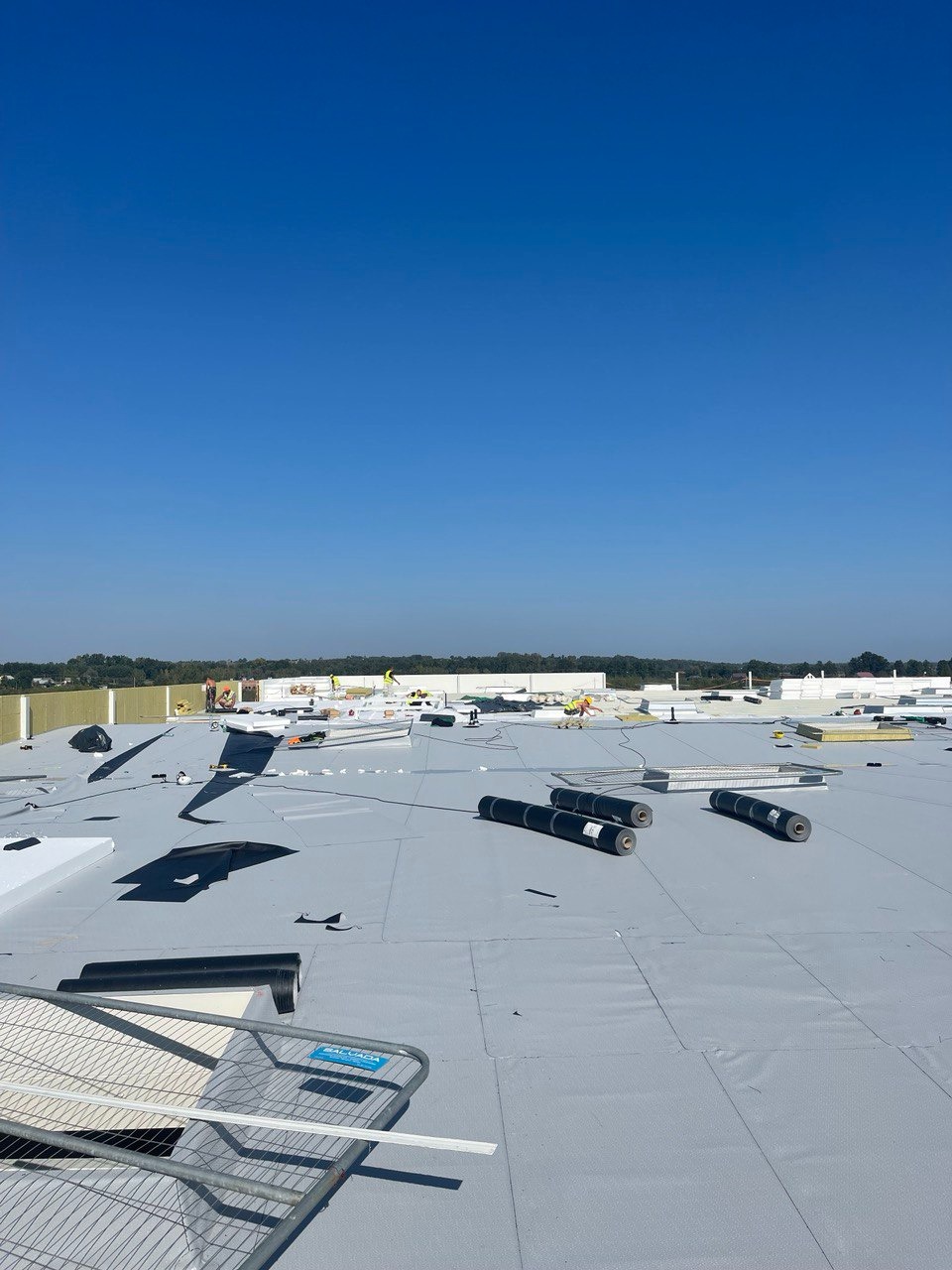 Rozległy, płaski dach pokryty szarą membraną z widocznymi rolkami materiału izolacyjnego, fragmentami białych płyt izolacyjnych i ekipą pracowników w oddali, na tle błękitnego nieba.