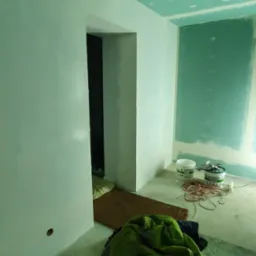Pokój w trakcie remontu: ściany i sufit częściowo wyszpachlowane i pomalowane na zielono, widoczne puszki z farbą, kabel elektryczny i robocze ubrania.
