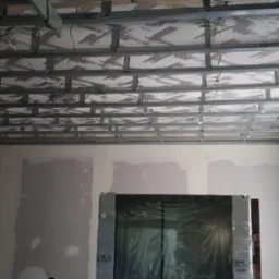 Metalowa konstrukcja sufitu podwieszanego z izolacją, widoczne ślady szpachlowania na ścianach w trakcie remontu.