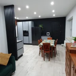 Jasne panele podłogowe w nowoczesnym salonie z aneksem kuchennym, widoczne szare szafki kuchenne, czarna ściana i drzwi, stół z krzesłami oraz zielona sofa z żółtą poduszką.