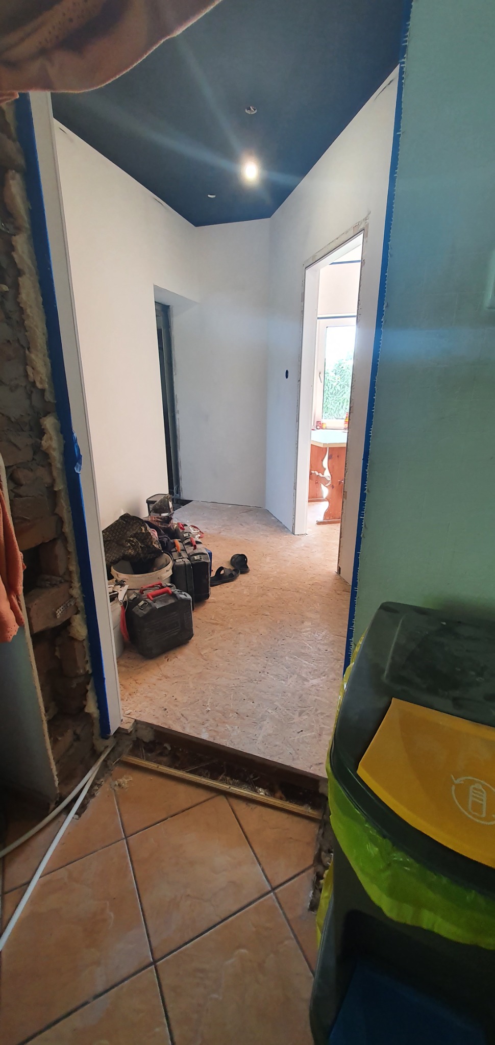 Wnętrze mieszkania w trakcie prac: pomalowany na ciemny kolor sufit, ściany przygotowane do malowania, widoczne narzędzia i materiały budowlane, podłoga częściowo wyłożona płytami OSB, częściowo...