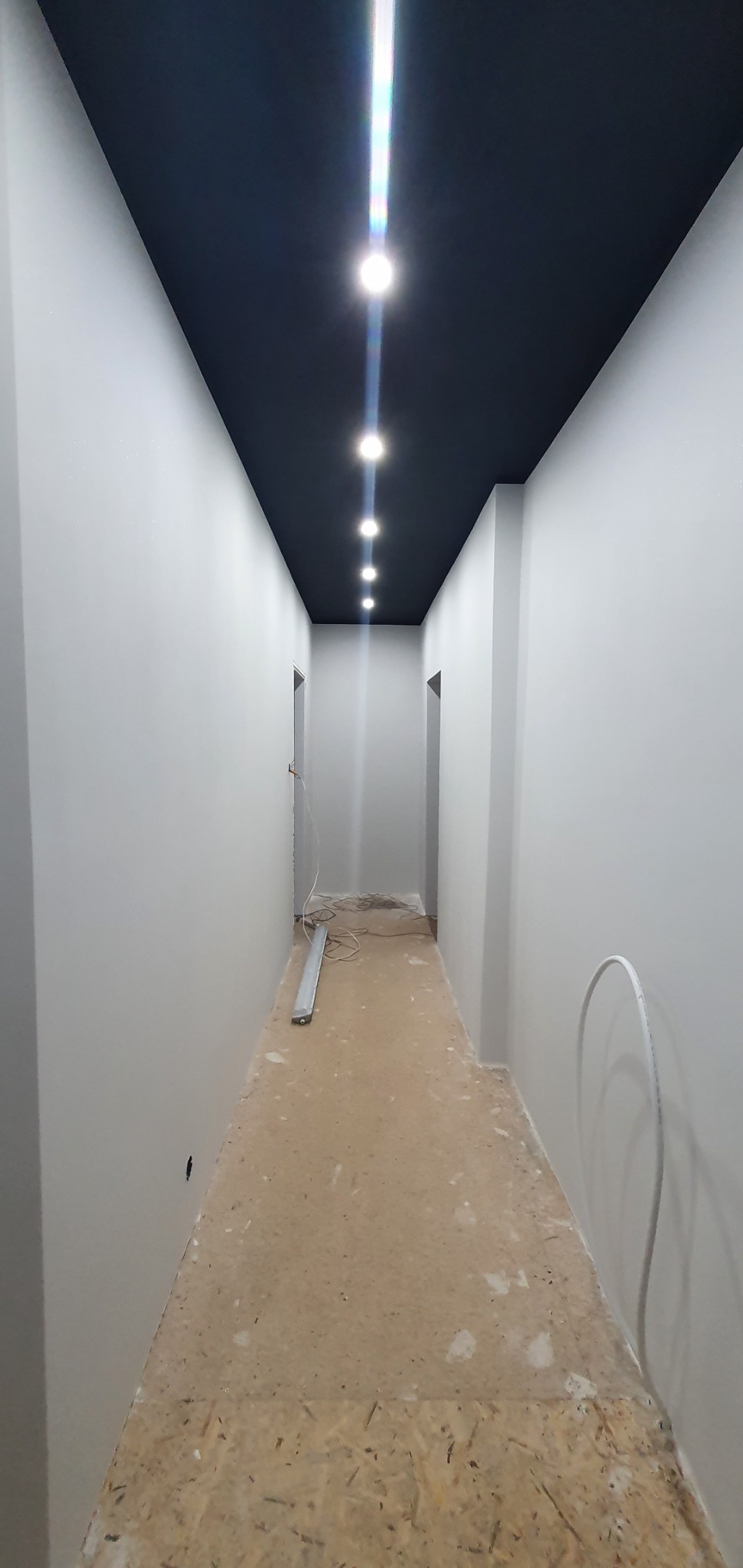 Korytarz w trakcie wykańczania z pomalowanymi na szaro ścianami i czarnym sufitem z rzędem punktowych lamp LED, widoczne kable i listwy przypodłogowe na podłodze.
