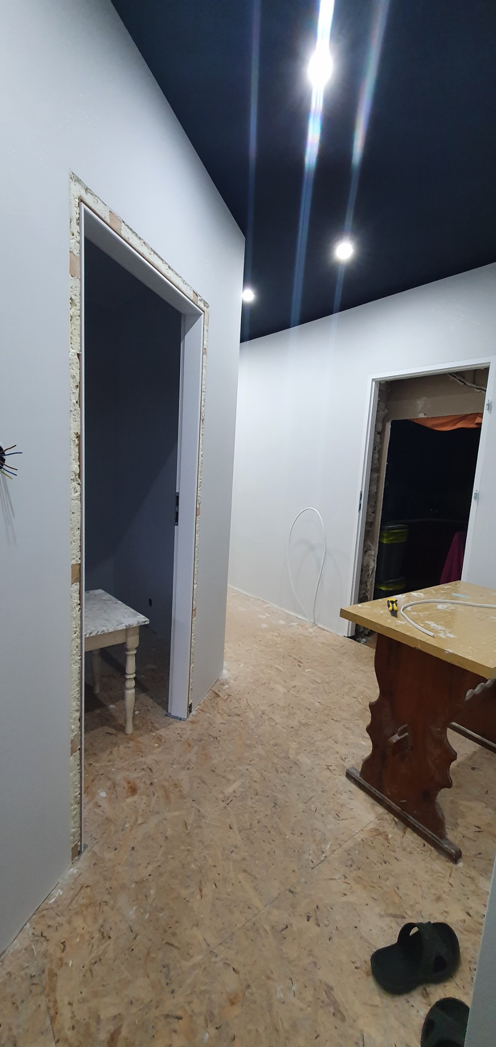 Wnętrze w trakcie prac wykończeniowych: surowa podłoga OSB, świeżo osadzone ościeżnice z widoczną pianką montażową, białe ściany, ciemny sufit z oświetleniem punktowym, widoczny fragment stołu...