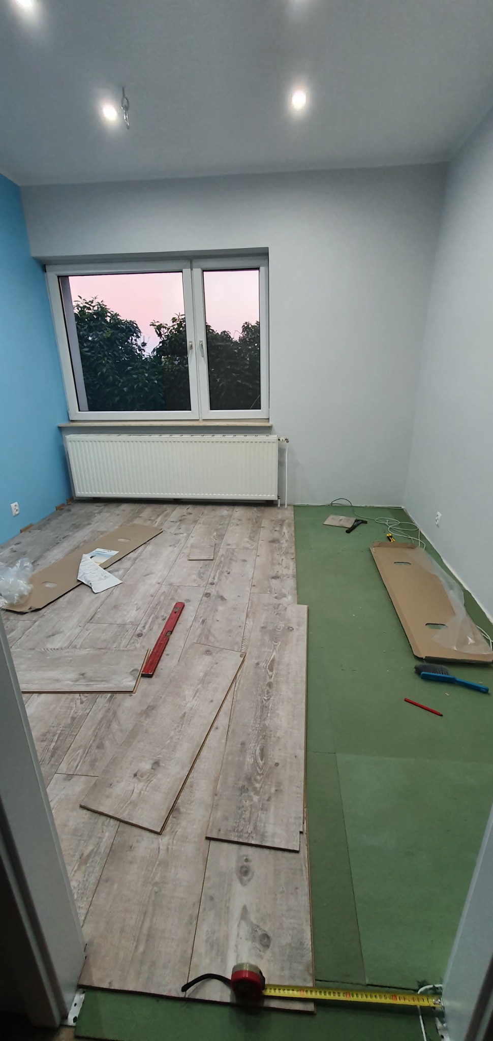Proces układania paneli podłogowych w pokoju w Chełmnie, widoczny fragment podkładu, rozłożone panele, poziomica i miara zwinięta przy listwie przypodłogowej.