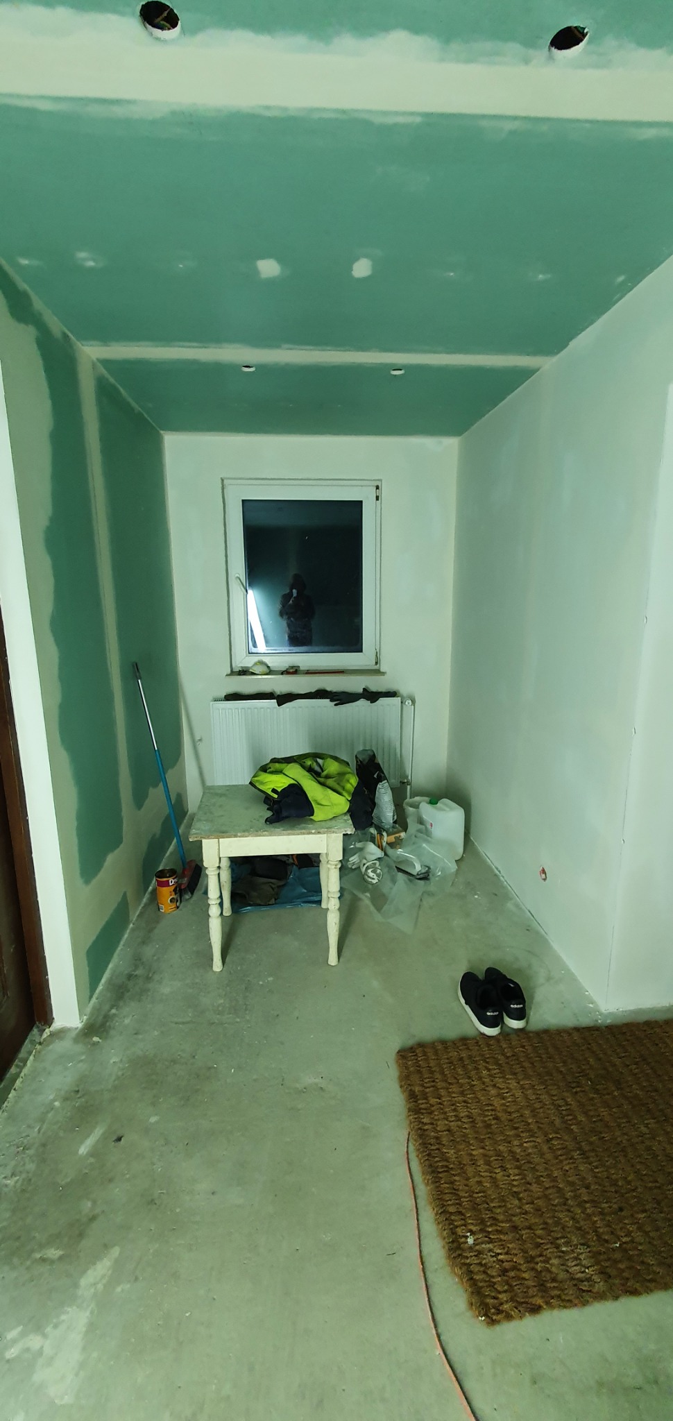 Pokój w trakcie prac wykończeniowych: ściany i sufit pokryte szpachlą, widoczne niedoskonałości i otwory na oświetlenie, na podłodze rozłożona mata, na stole robocza kurtka i narzędzia.